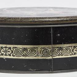 Side view of round black metal tin with lid. Central decorative gold band.