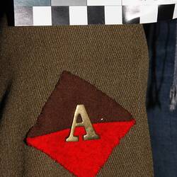 Rectangular brown and red colour patch with brass 'A' on khaki fabric.