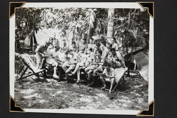 Group of men sitting on a table outside with trees and foliage behind.