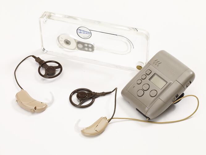 Rectangular grey coloured plastic battery pack with ear piece and circular magnetic processor attached by a long cord. The battery pack can attach to clothing by a clip on the back.