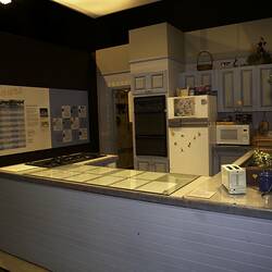 Kitchen studio set, pale blue bench. Contains kitchen appliances. Neighbours information panel on far wall.