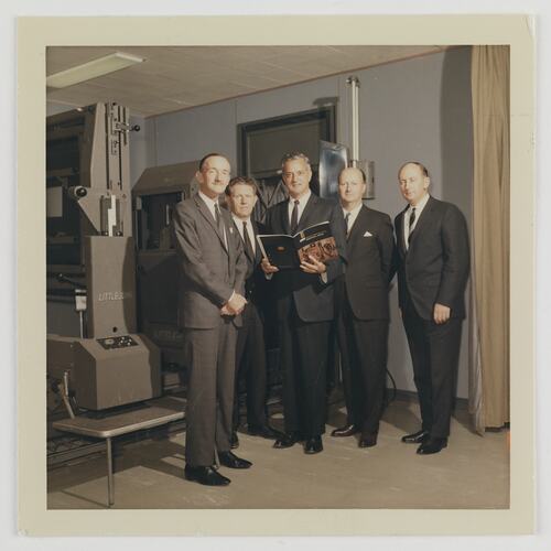 Five men wearing suits pose in front of a machine. The middle man holds an open catalogue.