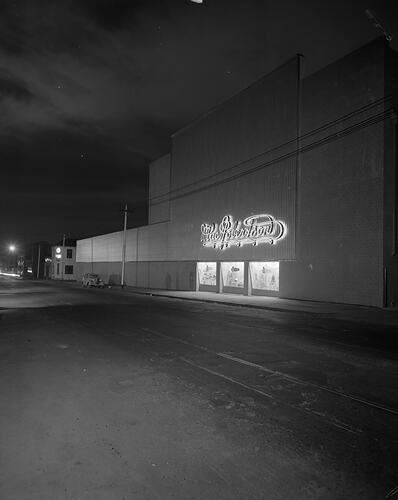 Building exterior of factory with three lit up windows. Neon sign above.