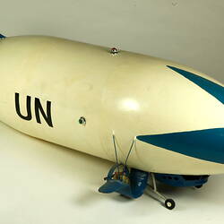 White model airship, blue nose and wings. Black UN on side. Small wheels at base. Front right view.