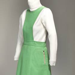 Angle view of lime green linen mini pinafore dress with zippered pockets on sides of skirt.