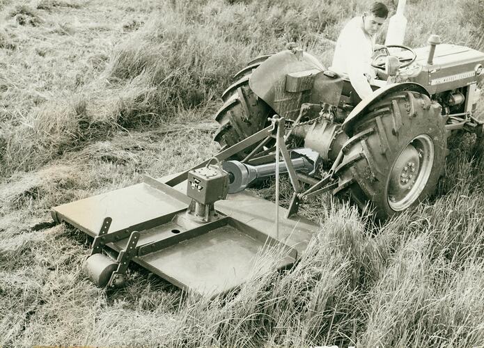 Man leaning over back of tractor seat, looking at a rotary slasher attached to back of tractor.