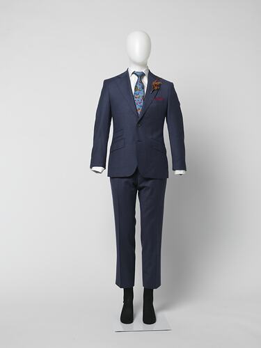 Blue suit jacket and pants with thin red square check pattern. Three outer pockets. Branded brown plastic buttons. White shirt, blue tie. Brown feather corsage.
