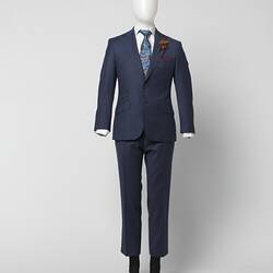 Blue suit jacket and pants with thin red square check pattern. Three outer pockets. Branded brown plastic buttons. White shirt, blue tie. Brown feather corsage.
