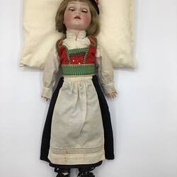 Doll with red, green, white top. White apron over black skirt. Red hat, black boots. Brown hair, eyes closed.