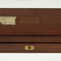 Closed wooden box with paper label on lid. Engraved brass plate inlaid in centre of lid.