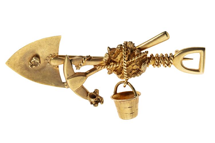 Gold brooch with pointed shovel, pick and bucket bound with rope around a larger nugget in centre. Small nugget on shovel and pick end.