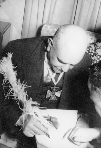 Man wearing a cross medal signing a document using a feather quill pen.