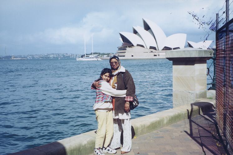 Woman and girl pose in front of Sydney Opera House building. Water behind them.