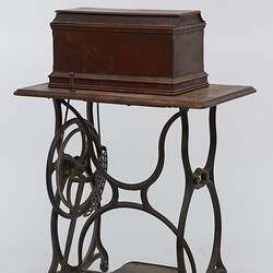 Sewing machine in wooden box fitted to a metal frame with a wooden bench and cover.