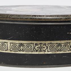 Side view of round black metal tin with lid. Central decorative gold band.