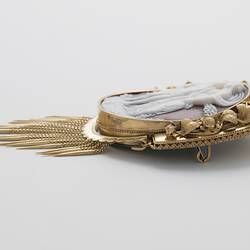 Oval cameo brooch with white winged figure and female, brown background. Ornate gold border and hanging fringe. Profile.