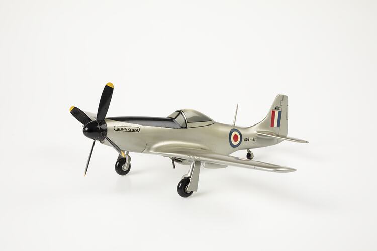 Wood model plane painted silver. Blue, white, red roundel on sides and wings. Red, white, blue flag on tail. Front black four blade propeller. Three wheels.