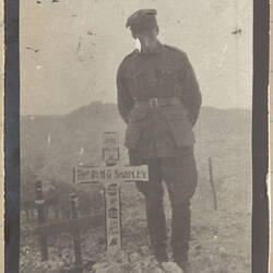 Soldier standing next to grave marked with white cross, other wooden crosses in background.
