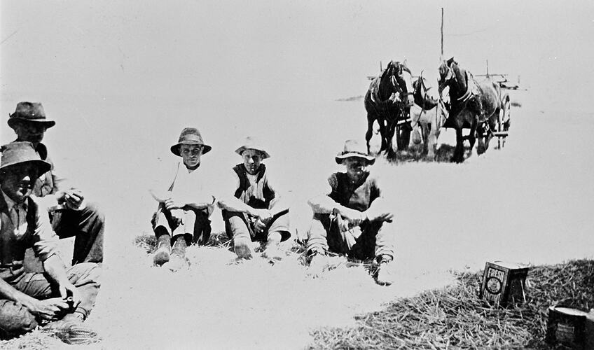 Five workmen sitting on the ground with a horse team in the background.