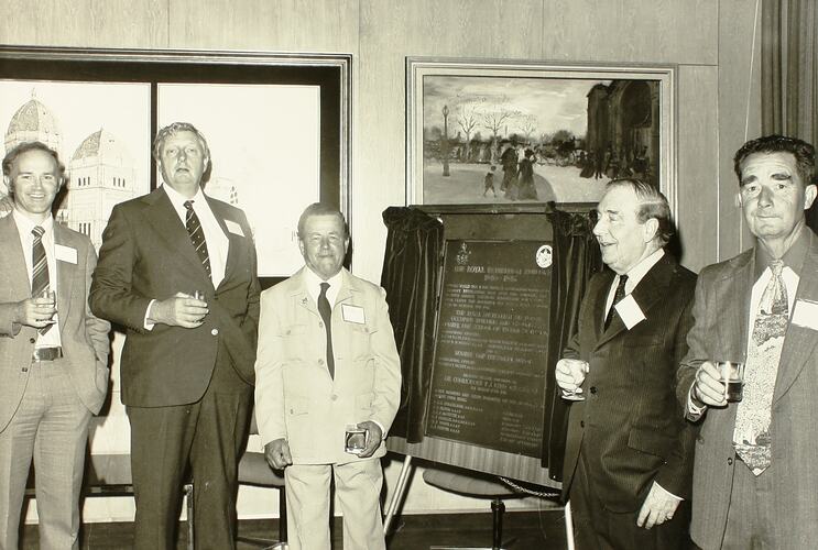 Black and white image of five men in suits, holding drinks in a room with framed paintings on the wall.