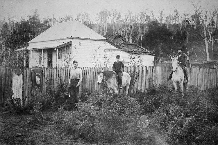 Man stands by fence at left wearing a glengarry. There is a sporran (and kilt?) hanging on the fence. Man and boy on horses at right.