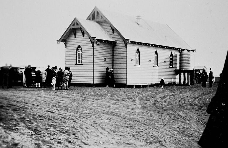 White weatherboard Lutheran church. Crowd out the front to the left. Water tank at right. Bare dirt foreground