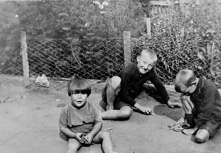 Three small boys sitting on the ground playing with wooden clothes pegs.