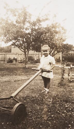 Infant girl in dress, pushing manual lawnmower. Garden, a large tree, and rocking horse in background.