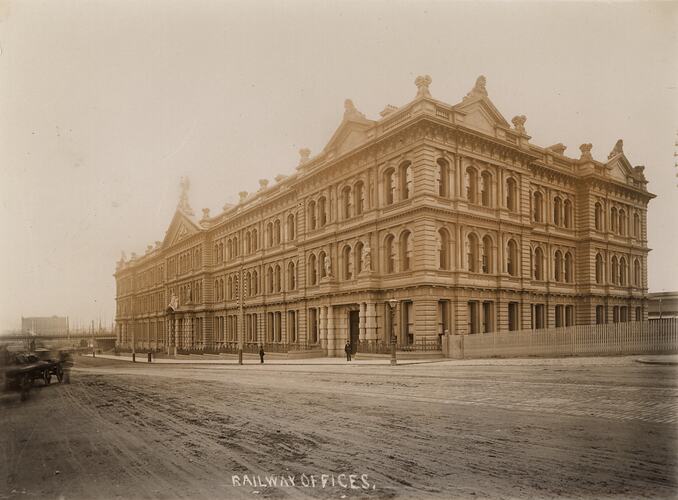 Corner view of highly decorated 3 storey stone building. Railway bridge, moored sailing ships in background.