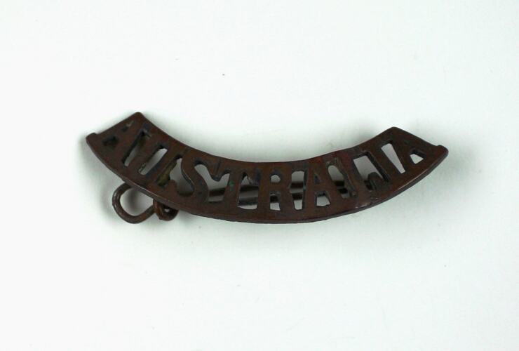 Bronze coloured metal badge in curved shape with word 'AUSTRALIA' cut out of metal.