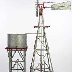 Model water pumping windmill next to water tank on a stand. Vanes side on facing left.