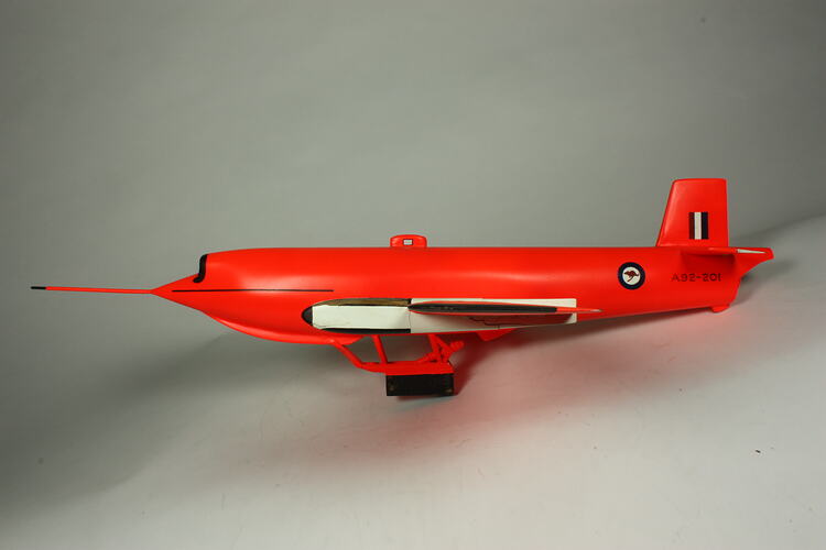 Orange pilotless aeroplane model with antenna attached to nose and a single skid in place of the wheels.