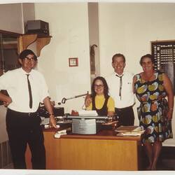 Two men and two women around a desk in an office. There is a typewriter in the desk they surround.