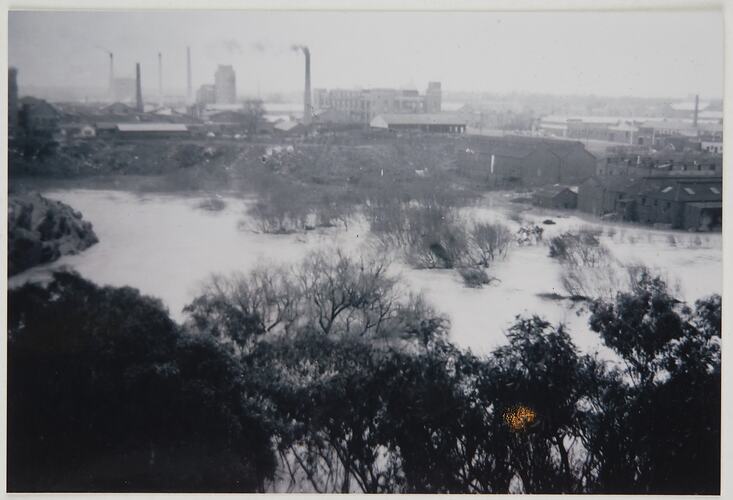Looking down over trees at flooded river in middleground. Buildings and factories on other side, partially sub
