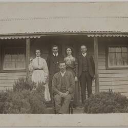 Group in front of weatherboard house with verandah. Two women and two men standing, one man sitting on front.
