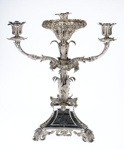 Ornate silver candelabra with three arms and central bowl. Decorative body sits on a tri-form base with three engraved plate.