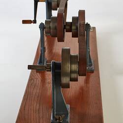 Wooden base with two pairs of round wheels, each connected via a pulley looped belt. One wheel has a handle.