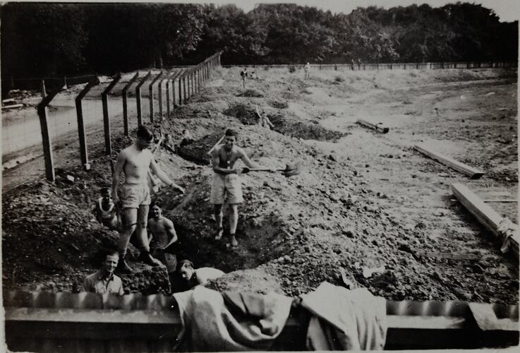 Six men in military shorts, mostly bare-chested, one wearing a shirt, digging a slit trench. Other men working on trenches in background.