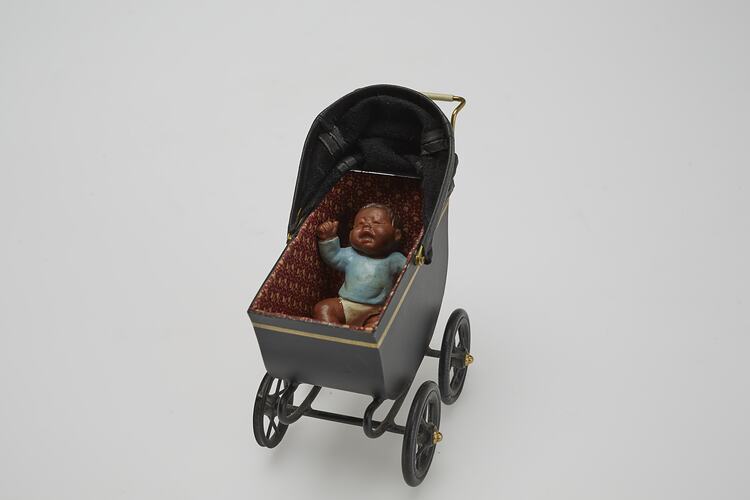 Black metal miniature pram, baby doll with dark brown hair and skin wearing blue top and white pants within.