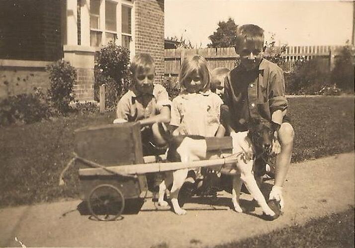 Young girl with three boys on footpath in front of house. Dog and cart in front of them.