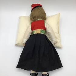 Back of doll with red and white top, gold belt and black skirt. Wears a red hat and black boots.