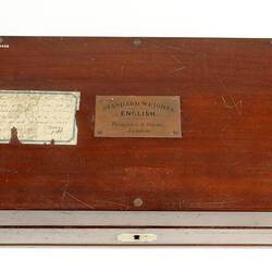 Closed wooden box with paper label on lid. Engraved brass plate inlaid in centre of lid.