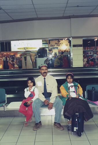 Man and two girls sitting in an airport with a suitcase.
