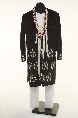 Black coat with white floral detail over white chinos pants. Long beaded necklaces.