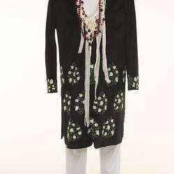 Black coat with white floral detail over white chinos pants. Long beaded necklaces.