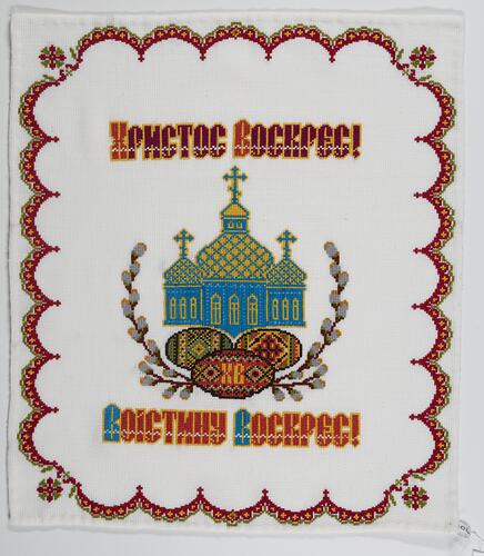 White cotton cloth with red, black yellow, green embroidered patterns and text. Orthodox church in centre.