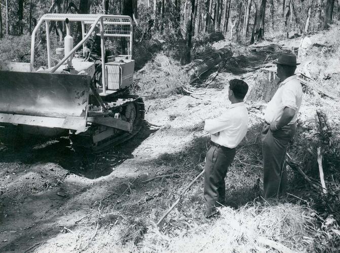 Two men watching a man driving a crawler tractor hauling a log on a cable.