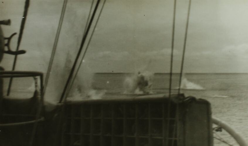 Multiple explosions in water, part of ship in foreground.