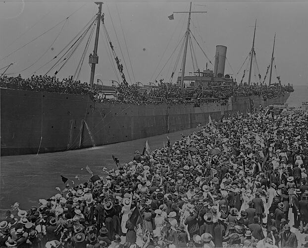 Large crowd on a wharf waving farewell to a troopship leaving a jetty.