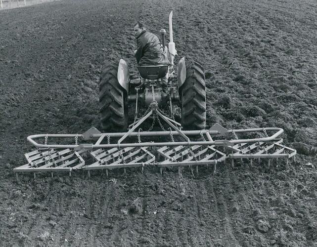 A man driving a tractor connected to a harrow, in a ploughed field.